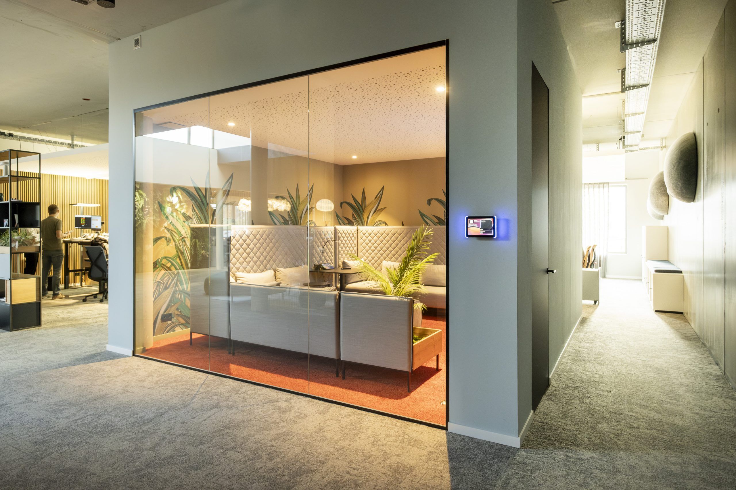 Kleiner Konferenzraum mit großen Fenstern, einem großen Tisch und gemütlichen Stühlen in blau mit blauem Teppich, im Hintergrund ein Fernseher, an der Wand ein sehr großes Bild mit Schubkarren vor Klinkermauer