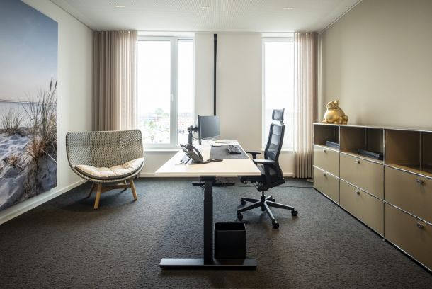 Heller Büroraum mit Schreibtisch zentral stehend auf dunklem Teppichboden. Am linken Rand ein Sessel vor Gardine, an der Wand ein großes Bild mit Strandmotiv. An der linken Seite ein braunes Sideboard mit goldenem Frosch darauf.